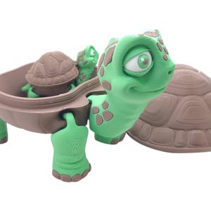 May include: A green turtle toy with brown spots and a brown shell. The turtle has a large, expressive face with a wide smile. The turtle is holding a smaller brown turtle toy inside its shell.