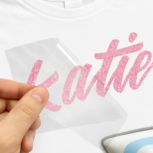 May include: A white t-shirt with the name "Katie" in pink glitter lettering. The glitter design is on a clear transfer sheet, held up to the shirt. A heat press is visible in the bottom right corner.
