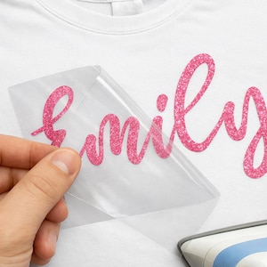 May include: A white t-shirt with the word "family" in pink glitter lettering. A clear transfer sheet is being peeled back, revealing the glitter design. An iron is visible in the bottom right corner.