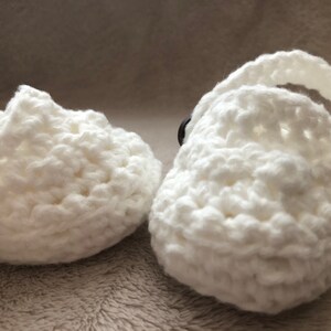 Peut inclure: Une paire de chaussons pour b&eacute;b&eacute; en crochet blanc avec des boutons noirs. Les chaussures ont un design simple et sont parfaites pour un nouveau-n&eacute;.