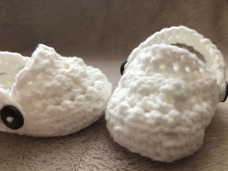 Peut inclure: Une paire de chaussons pour b&eacute;b&eacute; en crochet blanc avec des boutons noirs. Les chaussures ont un design simple et sont parfaites pour un nouveau-n&eacute;.