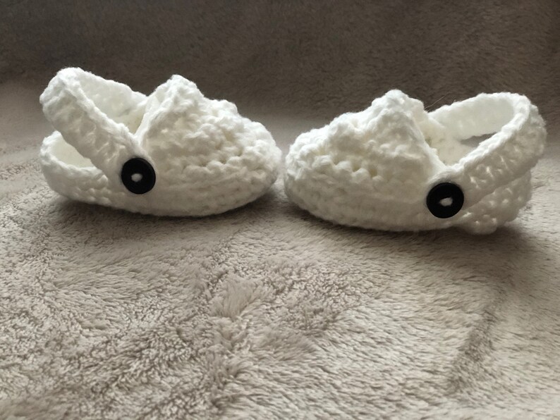 Peut inclure: Une paire de chaussons pour b&eacute;b&eacute; en crochet blanc avec des boutons noirs. Les chaussures ont une sangle qui passe autour de la cheville.