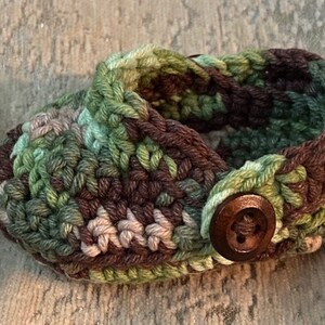 Peut inclure: Une paire de chaussons de b&eacute;b&eacute; en crochet avec un motif camouflage et un bouton marron. Les chaussons sont faits de fil vert, marron et beige.