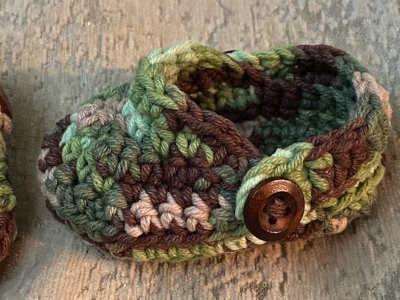 Peut inclure: Une paire de chaussons de b&eacute;b&eacute; en crochet avec un motif camouflage et un bouton marron. Les chaussons sont faits de fil vert, marron et beige.