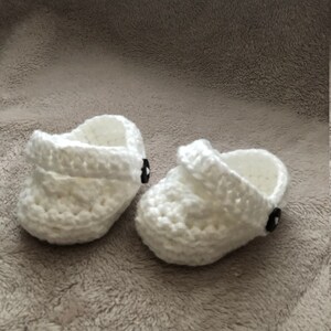 Peut inclure: Une paire de chaussons pour b&eacute;b&eacute; en crochet blanc avec des boutons noirs. Les chaussures ont un design simple et sont parfaites pour un nouveau-n&eacute;.