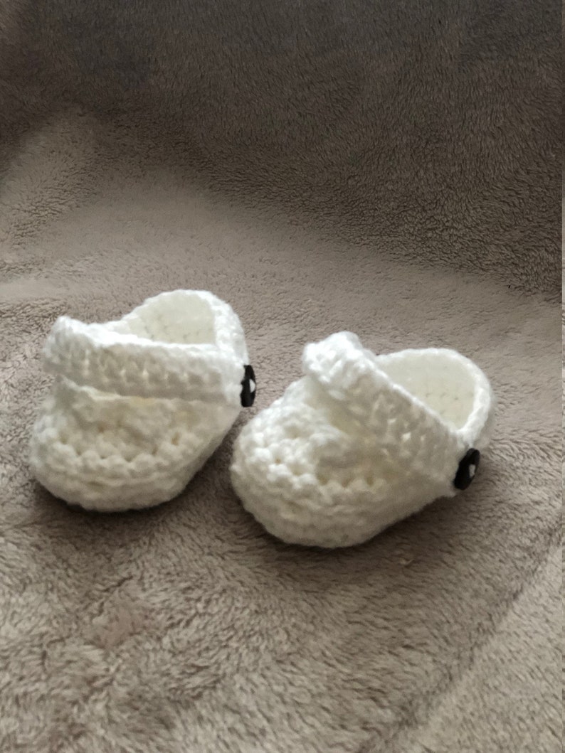 Peut inclure: Une paire de chaussons pour b&eacute;b&eacute; en crochet blanc avec des boutons noirs. Les chaussures ont un design simple et sont parfaites pour un nouveau-n&eacute;.