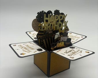 Könnte beinhalten: Eine schwarze und goldene Pop-up-Karte mit einem 3D-Design, das einen Geburtstagskuchen, Ballons und einen Hut zeigt. Die Karte ist geöffnet und innen steht "Happy Birthday".
