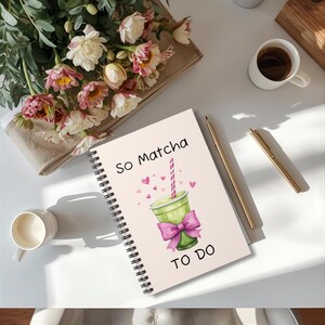 Op de afbeelding: Een spiraalgebonden notitieboek met de tekst "So Matcha" en "TO DO" op de kaft. De kaft toont een groene matcha-drank met een roze strik en hartjes. Een gouden pen en een kopje koffie staan ook op het witte oppervlak.