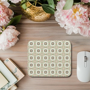 Può includere: Un tappetino per mouse rettangolare con un motivo floreale ripetuto nei toni del verde, beige e rosa. Un mouse per computer bianco è a destra del tappetino. Lo sfondo è una superficie in legno con fiori e libri.