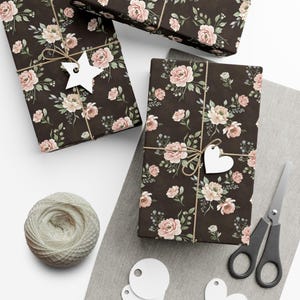 Op de afbeelding: Cadeauboxen verpakt met bloemmotiefpapier in roze en crème op een donkerbruine achtergrond. Elke doos is vastgebonden met touw en versierd met een witte ster- of hartvormige tag. Een bol touw, een schaar en blanco tags zijn ook aanwezig.