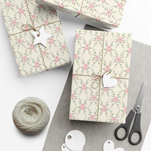 Op de afbeelding: Verschillende ingepakte cadeaus met een bloemenrozenpatroon in roze en groen op een crèmekleurige achtergrond. De cadeaus zijn vastgebonden met touw en hebben witte papieren labels. Een bol touw, een schaar en blanco labels zijn ook aanwezig.