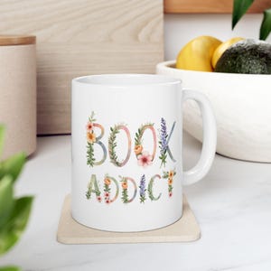 Op de afbeelding: Witte keramische mok met de tekst "BOOK ADDICT" in bloemrijke letters. De letters zijn versierd met aquarelbloemen en groen in roze, oranje en paarse tinten. De mok staat op een beige onderzetter.