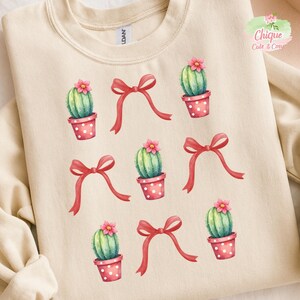 Op de afbeelding: Crèmekleurige sweatshirt met een herhaald patroon van cactussen in roze polkadot potten, rode strikken en roze bloemen. De cactussen zijn groen met roze bloemen. De sweatshirt heeft een ronde hals.