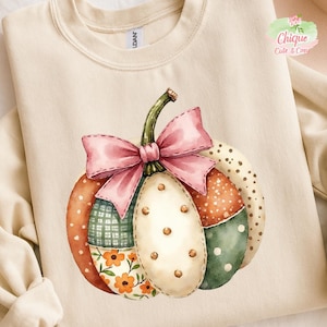 Op de afbeelding: Crèmekleurige sweatshirt met een aquarel pompoenontwerp. De pompoen is samengesteld uit verschillende stofpatronen, waaronder bloemen, stippen en ruiten, en is bekroond met een roze strik. Het ontwerp is gecentreerd op de voorkant van het shirt.