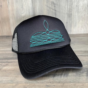 May include: A black trucker hat featuring a structured foam front panel and a breathable mesh back. The front panel displays an embroidered teal graphic consisting of horizontal lines and a central decorative flourish pattern in a stitch design.