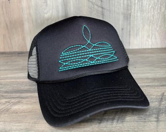 May include: A black trucker hat featuring a structured foam front panel and a breathable mesh back. The front panel displays an embroidered teal graphic consisting of horizontal lines and a central decorative flourish pattern in a stitch design.