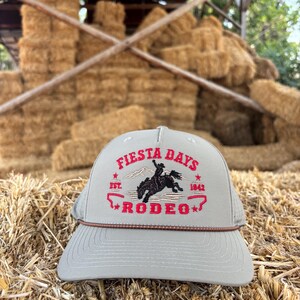 May include: A light gray baseball cap featuring a brown rope detail across the base of the crown. The front displays red embroidered text reading FIESTA DAYS RODEO, with EST. 1842 and a black silhouette of a bucking horse and rider.