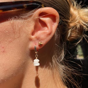May include: A close-up of a person's ear adorned with two earrings. One is a small gold star stud, and the other is a gold hoop with three stacked white beads. The person is wearing tortoise shell sunglasses.