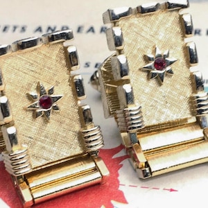 May include: Gold-coloured cufflinks with a textured rectangular design. Each cufflink features a starburst design with a red gemstone in the centre. The cufflinks are set against a vintage map background.