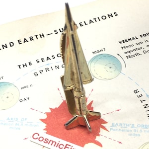 May include: A gold-colored metal tie clip in the shape of a rocket. The clip is set against a vintage map with text that includes "Earth-Sun Relations" and "CosmicFirefly". The rocket has a pointed nose and fin-like structures.