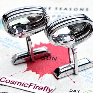 May include: Silver cufflinks with a geometric design. Each cufflink has a central, faceted, grey gemstone and a rectangular post. The cufflinks are set against a vintage map background, with the text "CosmicFirefly" visible.