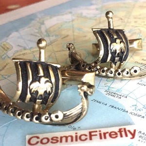 May include: Two gold-coloured Viking longboat cufflinks. Each cufflink has a black and gold striped sail with a detailed emblem. The cufflinks are presented on a map background, with the text "Cosmic Firefly" visible.