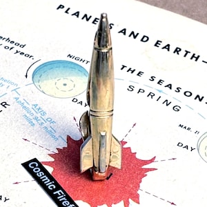 May include: A gold-coloured rocket-shaped lapel pin. The pin is set against a vintage map with text that reads "PLANES AND EARTH" and "Cosmic Firefly". The pin has a detailed design with fins and a pointed top.