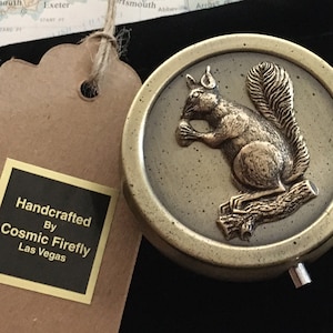 May include: A round, antique-style metal compact with a detailed relief of a squirrel holding a nut, perched on a branch. The compact is in a black velvet pouch, with a tag that reads "Handcrafted by Cosmic Firefly, Las Vegas."