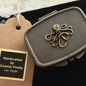 Puede incluir: Una caja rectangular de metal gris con tapa abisagrada, adornada con un emblema de pulpo de latón. La caja va acompañada de una bolsa de terciopelo negro y una etiqueta que dice "Handcrafted by Cosmic Firefly Las Vegas."