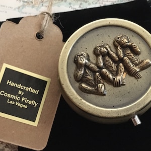 May include: A round, antique-style trinket box with a bronze finish. The lid features three monkey figures in the "see no evil, hear no evil, speak no evil" pose. The box is next to a tag that says "Handcrafted By Cosmic Firefly Las Vegas."