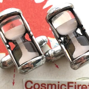May include: A pair of silver cufflinks, each with a small hourglass design. The hourglasses have a clear glass centre with white and red sand, set in a silver frame. The cufflinks are on a colourful background.