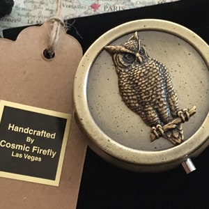 Puede incluir: Una caja de baratijas redonda de estilo antiguo con un búho de bronce detallado posado en una rama. La caja está colocada contra un mapa del mundo y una bolsa de terciopelo negro. La etiqueta dice "Handcrafted By Cosmic Firefly Las Vegas."