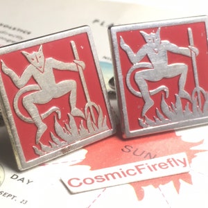 May include: Pair of square cufflinks with a red background and a silver devil figure holding a pitchfork. The devil is depicted above flames. The cufflinks have a silver border.
