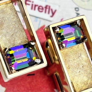 May include: Gold-coloured rectangular cufflinks featuring iridescent, multi-coloured rectangular crystals. The cufflinks have a textured gold-coloured centre and are set against a red and white background. The word "Firefly" is visible in the background.