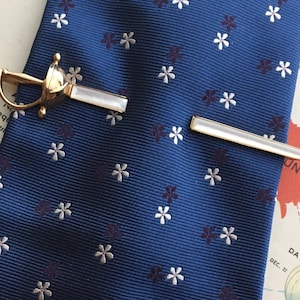 以下が含まれることがあります： 金色のネクタイクリップと、真珠母貝のアクセントが付いた剣型のネクタイバーが、花柄の紺色のネクタイの上に置かれています。 クラシックなアクセサリーです。