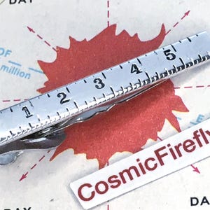 Könnte beinhalten: Eine silberfarbene Krawattenklammer in Form eines Lineals, mit Messungen bis zu 12,7 cm. Die Klammer befindet sich auf einem Kartenhintergrund mit einer roten Form und dem Text "CosmicFirefly".