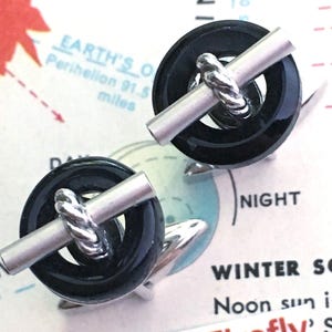 May include: A pair of cufflinks with a black, circular design, a silver-coloured bar, and a twisted silver-coloured detail. The cufflinks are set against a map background. The text "CosmicFirefly" is visible.
