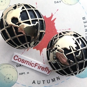 May include: A pair of globe-shaped stud earrings. The earrings have a black base with a gold-coloured world map design and a grid pattern. They are displayed on a vintage map with the text "CosmicFirefly".