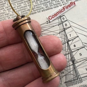 May include: A brass hourglass ornament with a gold cord. The hourglass is filled with white sand and is hanging from a gold cord. The background is a page from a book about sailing.