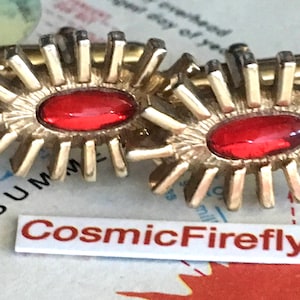 May include: A pair of vintage gold-tone clip-on earrings. Each earring has a central oval red stone and a sunburst design. The earrings are set against a map background, with the text "CosmicFirefly" visible.