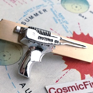 May include: A silver-colored toy ray gun with the text "Craftsman 100" on a light brown rectangular base. The ray gun has a detailed design and is set against a colorful background with text and graphics.