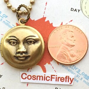 May include: A gold locket necklace with a detailed face of the moon on the front. The locket is hanging from a gold chain with small round beads. A penny is included in the image for size comparison.