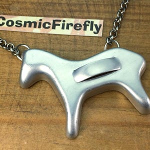 May include: Silver-toned metal pendant necklace featuring a stylized horse design. The pendant is attached to a silver chain. The necklace is displayed on a wooden surface. The text "CosmicFirefly" is visible in the background.