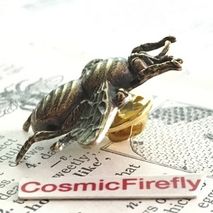 May include: A bronze-toned metal bee lapel pin featuring detailed wing textures and a segmented body. The pin includes a gold-colored butterfly clutch backing. A white rectangular label with the red text CosmicFirefly is positioned below.