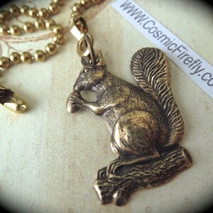 Op de afbeelding: Een gouden metalen eekhoornbeeldje met een borstelige staart, zittend op een boomstam, bevestigd aan een gouden ketting met een clip. De eekhoorn houdt een noot in zijn poten. De ketting wordt gebruikt om een ​​plafondventilator te trekken.