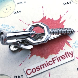 May include: A silver-coloured tie clip shaped like a bolt and a hex nut. The clip has a screw-like design on one end and a hexagonal shape in the middle. The clip is on a vintage map with the text "CosmicFirefly".