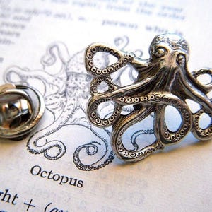 May include: Silver octopus pin with a detailed design. The pin is on a white background with a drawing of an octopus.