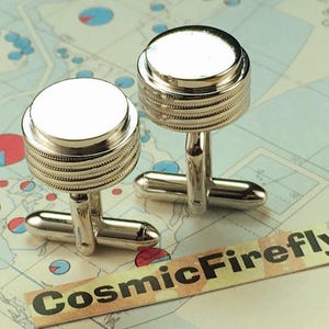 May include: A pair of silver cufflinks featuring a layered design and a white circular top. The cufflinks are presented against a map backdrop. The text "CosmicFirefly" is displayed on a small label.
