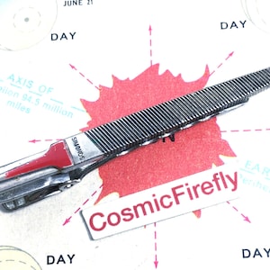 May include: A silver-toned tie clip with a textured, ridged design. The clip features a red accent and the brand name "SIMONDS". The background includes a map-like design with text and the words "CosmicFirefly".