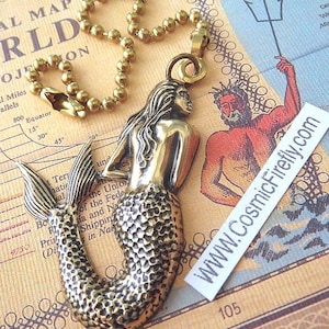 May include: A brass mermaid pendant on a beaded chain. The mermaid has detailed scales and flowing hair. The pendant is displayed on a vintage map with the text "WWW.CosmicFirefly.com".